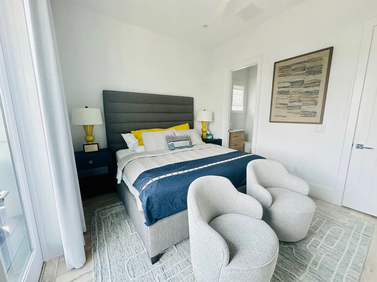 A bright, serene bedroom featuring a bed with a gray headboard, colorful yellow and blue bed linens, two modern chairs beside the bed, and framed artwork on the walls. Natural light pours in, emphasizing a peaceful and cozy vibe.