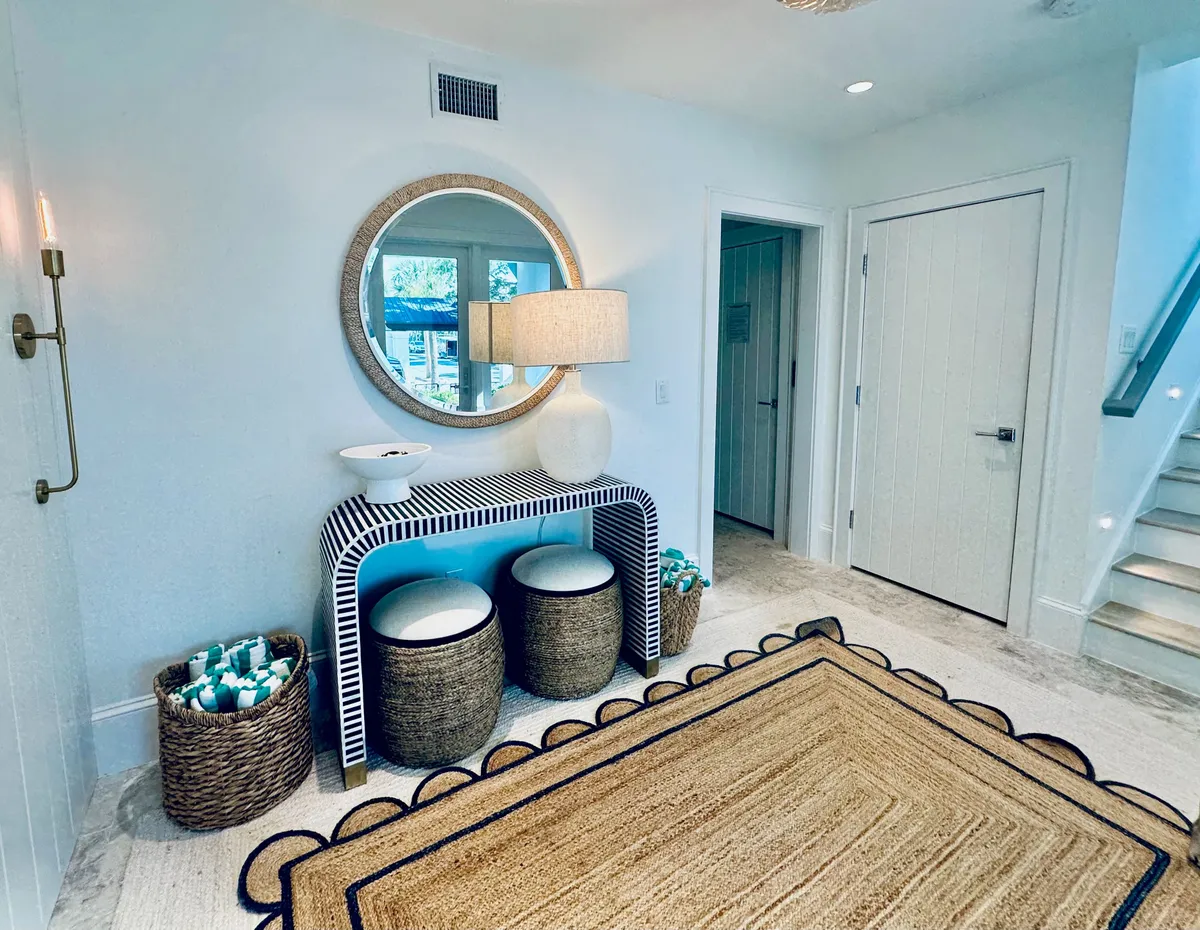 The image features a stylish entryway with a round mirror above a striped console table, a table lamp, and woven baskets on a rug, creating an inviting and modern ambiance. The vignette is open and bright, setting the tone for the home's interior.
