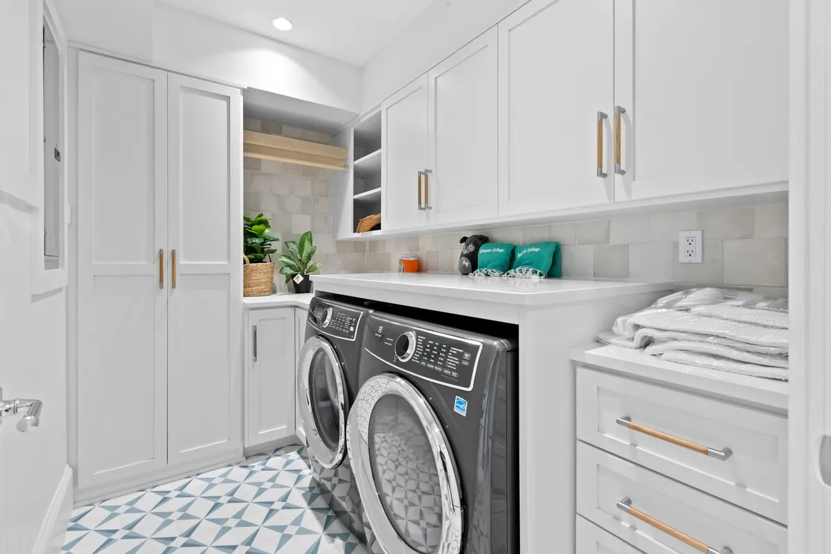 A functional laundry space with a washer and dryer, cabinetry, a decorative basket, and a green plant sits on textured floor tiles. The space is bright and organized, showcasing the home's commitment to accessibility and convenience for daily living.