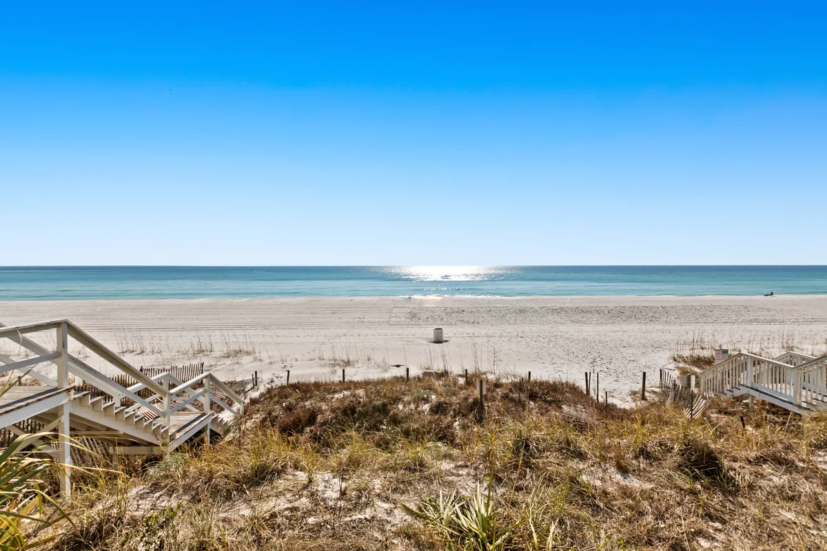 A sweeping view of the ocean and wide sandy beach is framed by dune grasses, with steps leading directly down to the soft sand. The tranquil shoreline is empty, offering a pristine, private-feeling stretch of coast, perfect for sunbathing, sandcastle building, or leisurely walks.