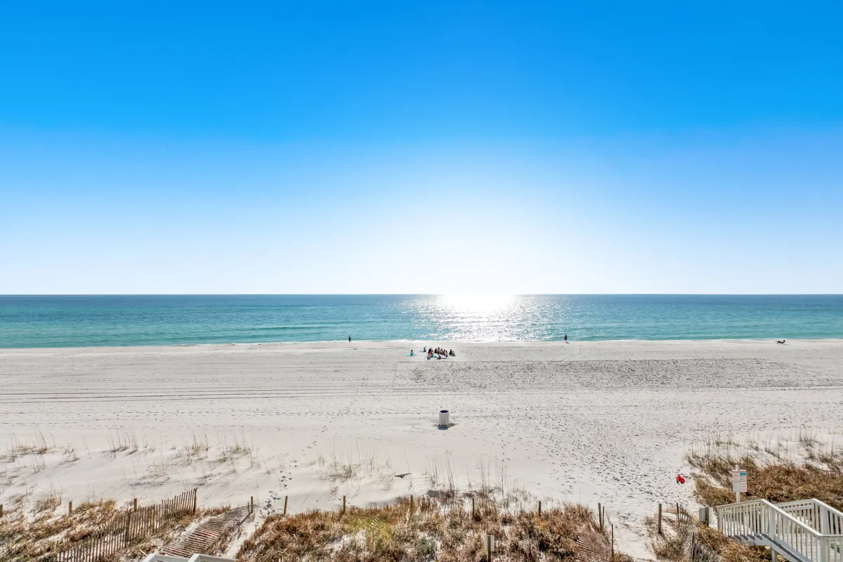 A panoramic perspective of a vast ocean and sunlit sandy beach, dotted with distant figures engaged in beach activities. The scene is bright and peaceful, capturing the vibrancy and fun of seaside adventures—ideal for families and beachgoers.