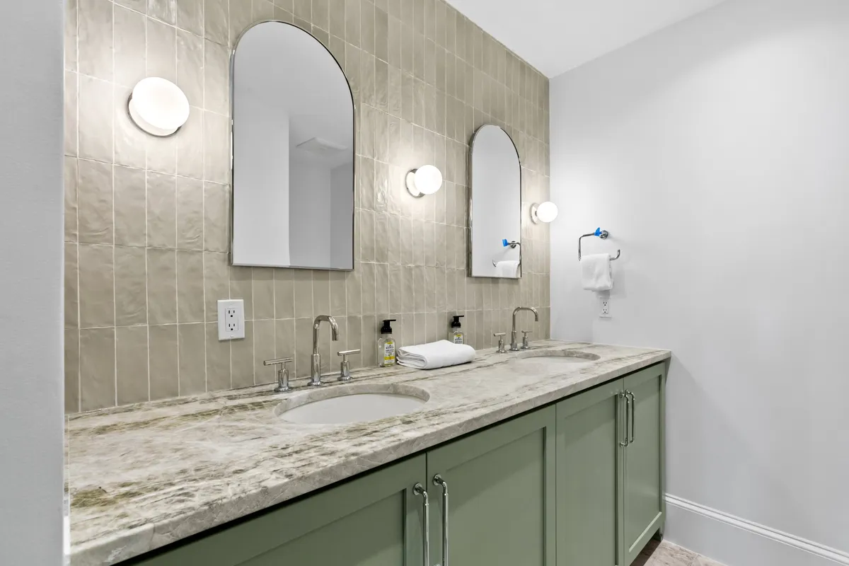 A stylish bathroom with dual sinks, arched mirrors, textured wall tiles, and green cabinetry. Morning sunlight reflects off the surfaces, emphasizing a fresh and inviting space ideal for personal routines and relaxation.