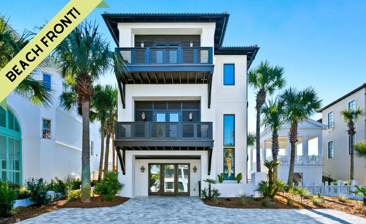 The image showcases a stunning three-story white coastal home with modern dark balcony railings, expansive glass doors and windows reflecting the surrounding palm trees and blue sky. The home's grand entrance is framed by tall palms, and a paved driveway leads guests to the front door, highlighting the elegance and serenity of the property. Visible upper balconies promise breathtaking, uninterrupted ocean views, and the beach is just steps away via private boardwalk access.