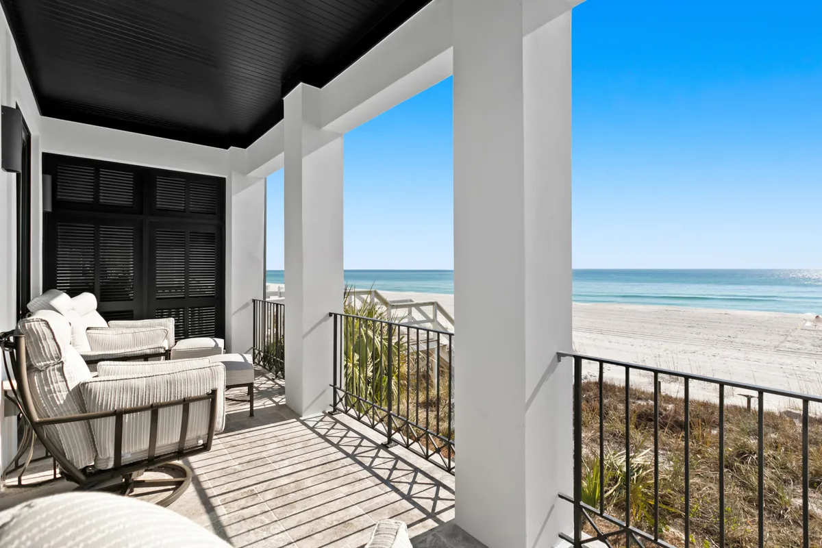 A beautifully furnished balcony with comfortable cushioned seating overlooks a wide-open ocean view. White architectural columns and black shutters frame the space, inviting guests to relax and enjoy the soothing sounds and sights of the Gulf. The tranquil setting is ideal for sunset gatherings, private toasts, and quiet conversations, all set against a panoramic coastal backdrop.