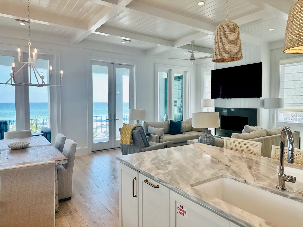 A spacious open-concept interior featuring a marble kitchen island with modern fixtures, a cozy living room with a large sectional sofa and mounted TV, and a dining area with a rustic wooden table and chandelier. Large glass doors frame breathtaking ocean views, filling the space with natural light and offering seamless indoor-outdoor flow.
