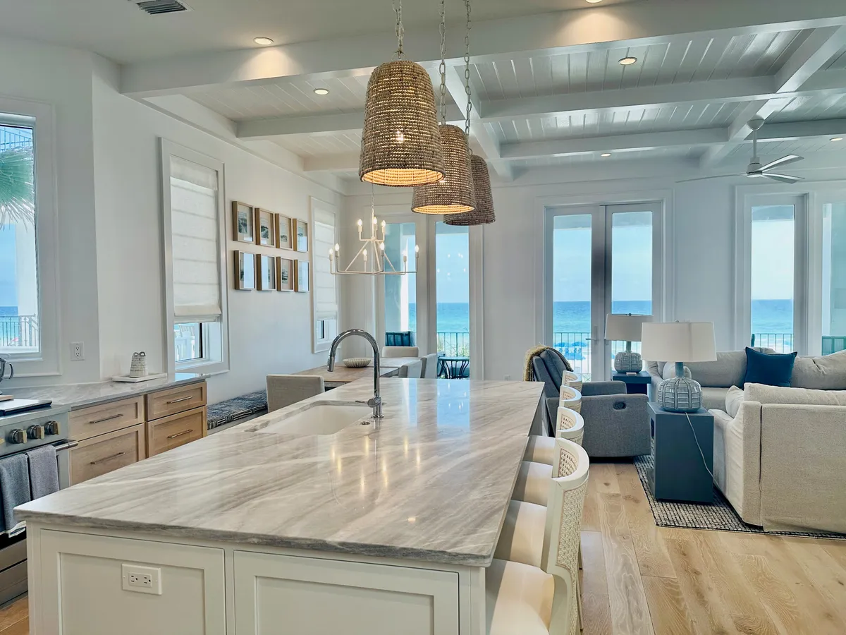 A bright, inviting interior with a large marble kitchen island, woven pendant lights, and a comfortable seating area oriented toward expansive floor-to-ceiling windows. These windows provide an unobstructed view of the beach and ocean, flooding the space with sunlight and connecting the interior to the coastal landscape.