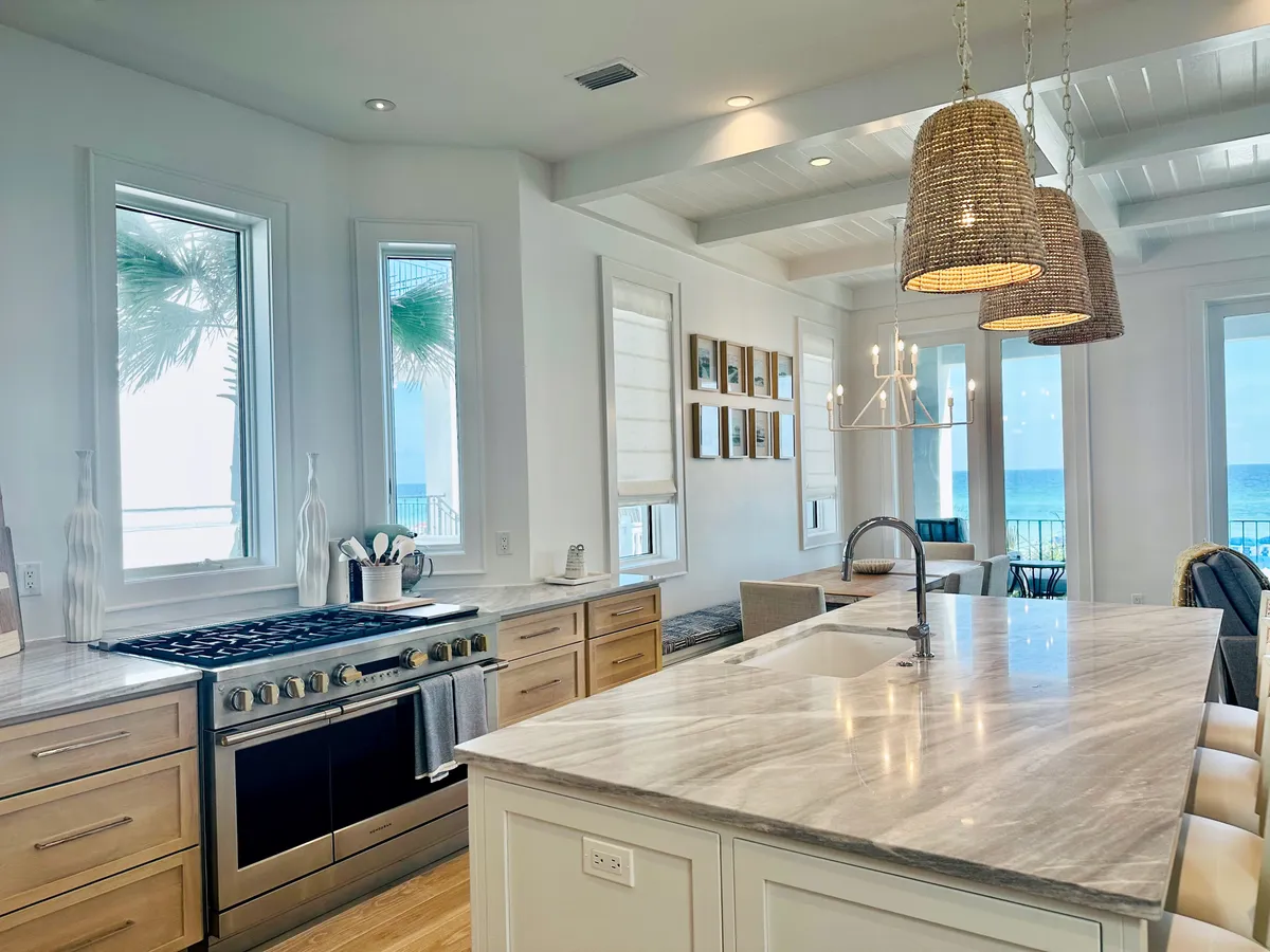 A luxurious kitchen featuring a marble island with an integrated sink, a modern gas stove with stainless steel finish, and large windows that frame stunning ocean views. Woven pendant lights hang above the island, and the space exudes upscale comfort and brightness.