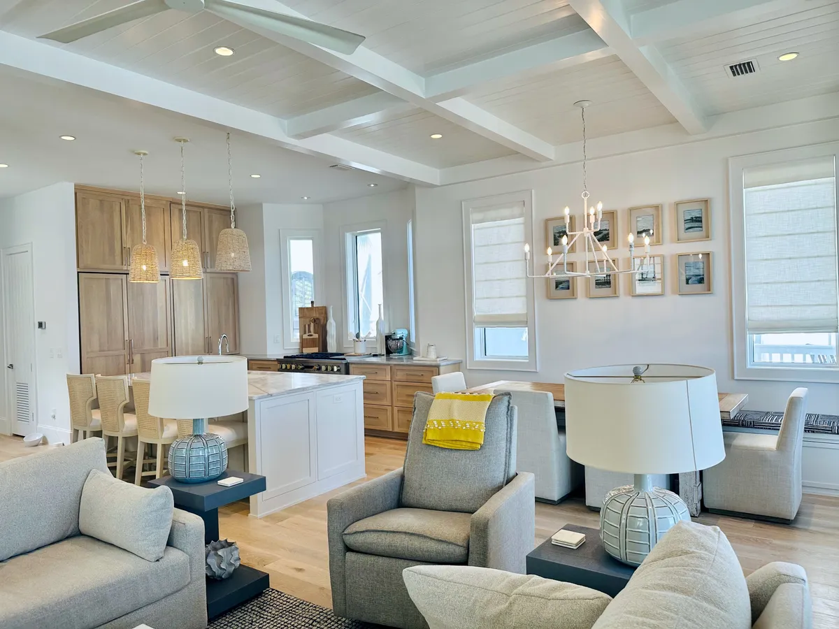 A sunlit interior displaying a cozy gray armchair with a yellow throw, a kitchen island with pendant lighting, and a dining area adorned with a chandelier and framed artwork. Large windows allow natural light to flood the space, creating a welcoming atmosphere that blends the kitchen and dining area.