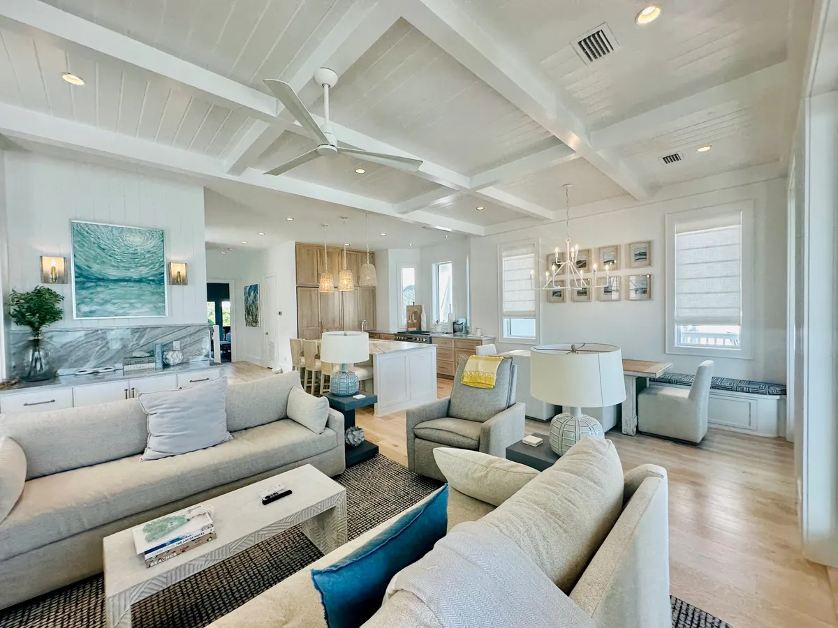 A spacious and elegant living room features comfortable grey sofas arranged for conversation, a modern kitchen in the background, and large windows showcasing coastal views. The setting is designed for socializing, hosting gourmet meals, or simply unwinding after a day outdoors, with seamless transitions between indoor comfort and outdoor allure.
