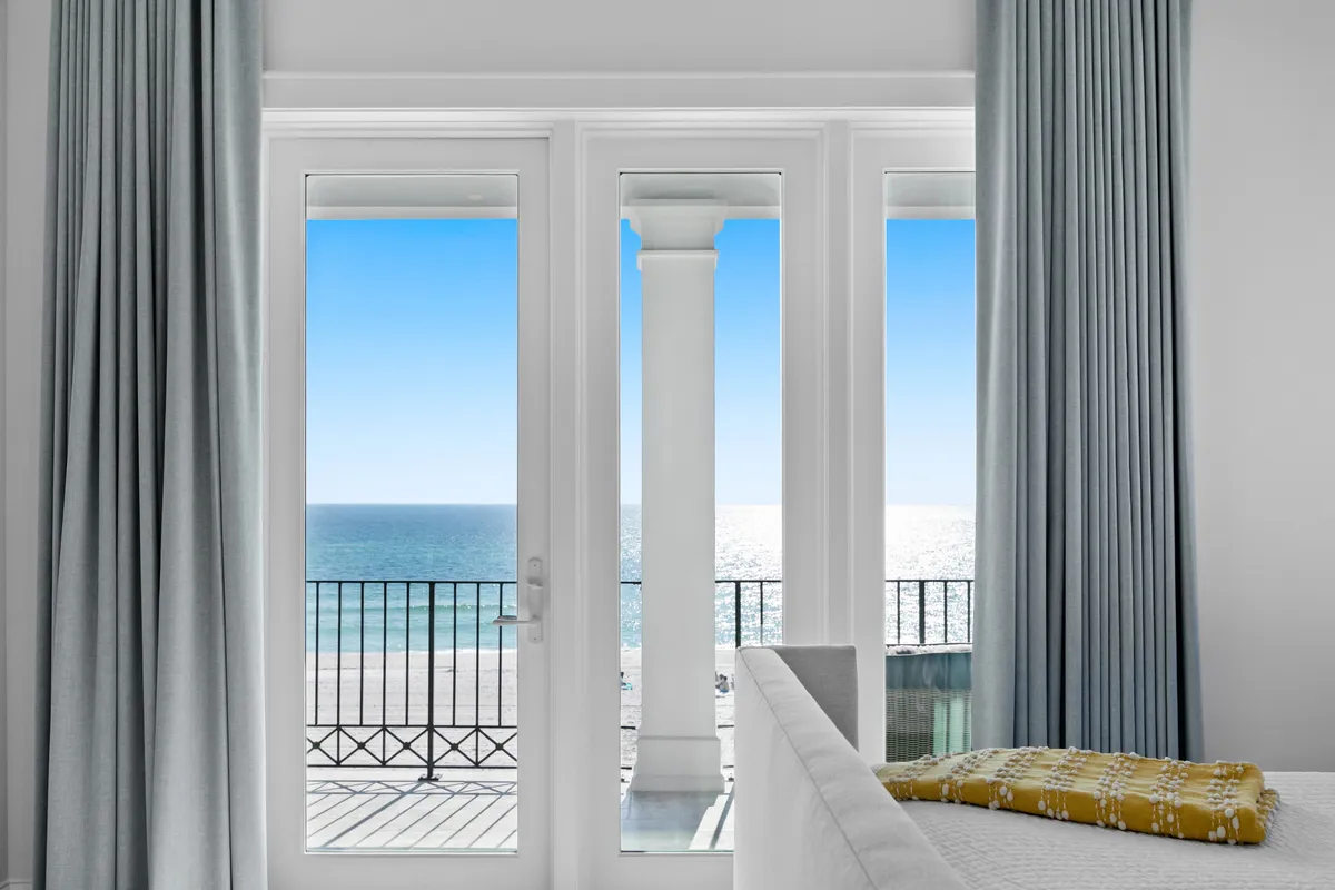 Inside a sunlit bedroom, French doors open directly onto a balcony with a stunning ocean view. A king bed with a yellow throw sits in the foreground, while white columns frame the blue horizon outside. Soft curtains add a touch of coastal luxury, reinforcing a tranquil and rejuvenating atmosphere.