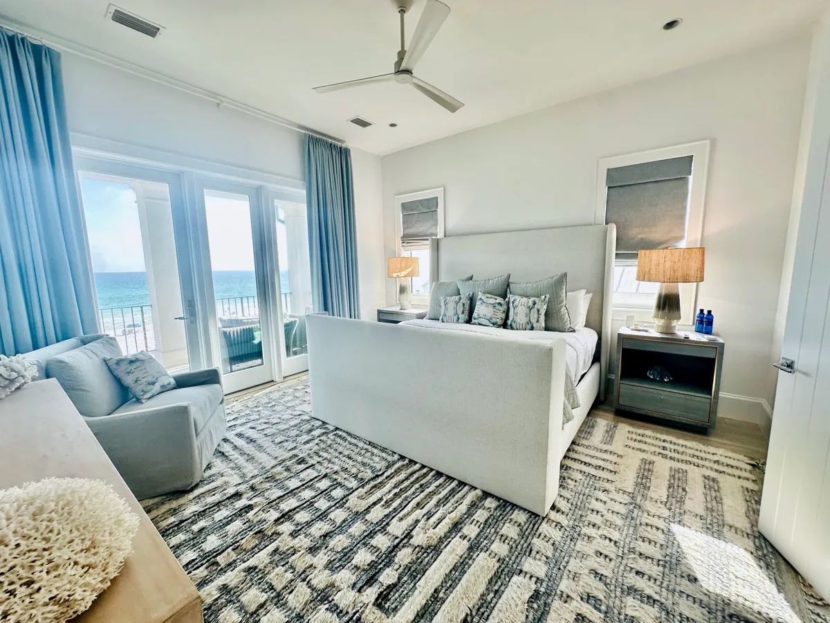 A spacious bedroom featuring a large upholstered bed adorned with decorative pillows, French doors that open to a private balcony offering ocean views, a plush armchair by the window, and a textured area rug. The soft natural light and serene decor create a tranquil and restful atmosphere.