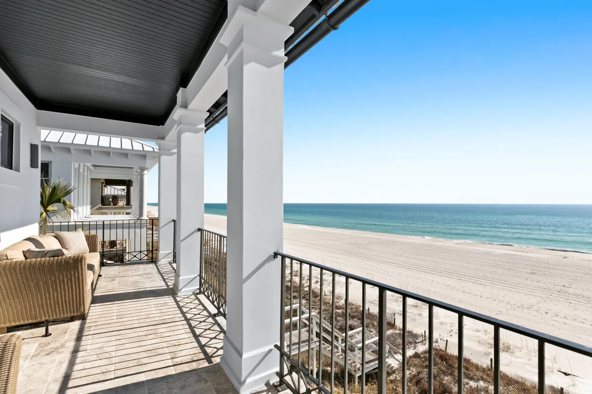 A wide porch with comfortable wicker seating overlooks an expansive sandy beach and calm turquoise ocean, framed by white columns and black railing. The private boardwalk is visible, indicating immediate access to the beach from the comfort of the home.