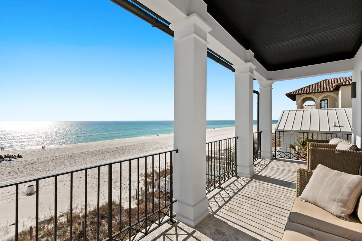 A spacious balcony with comfortable seating overlooks the sparkling ocean, framed by elegant white columns and black railings. The sandy beach stretches out below, offering uninterrupted views of the water and sky. The setting exudes calm and luxury, inviting relaxation and enjoyment of the sea breeze.