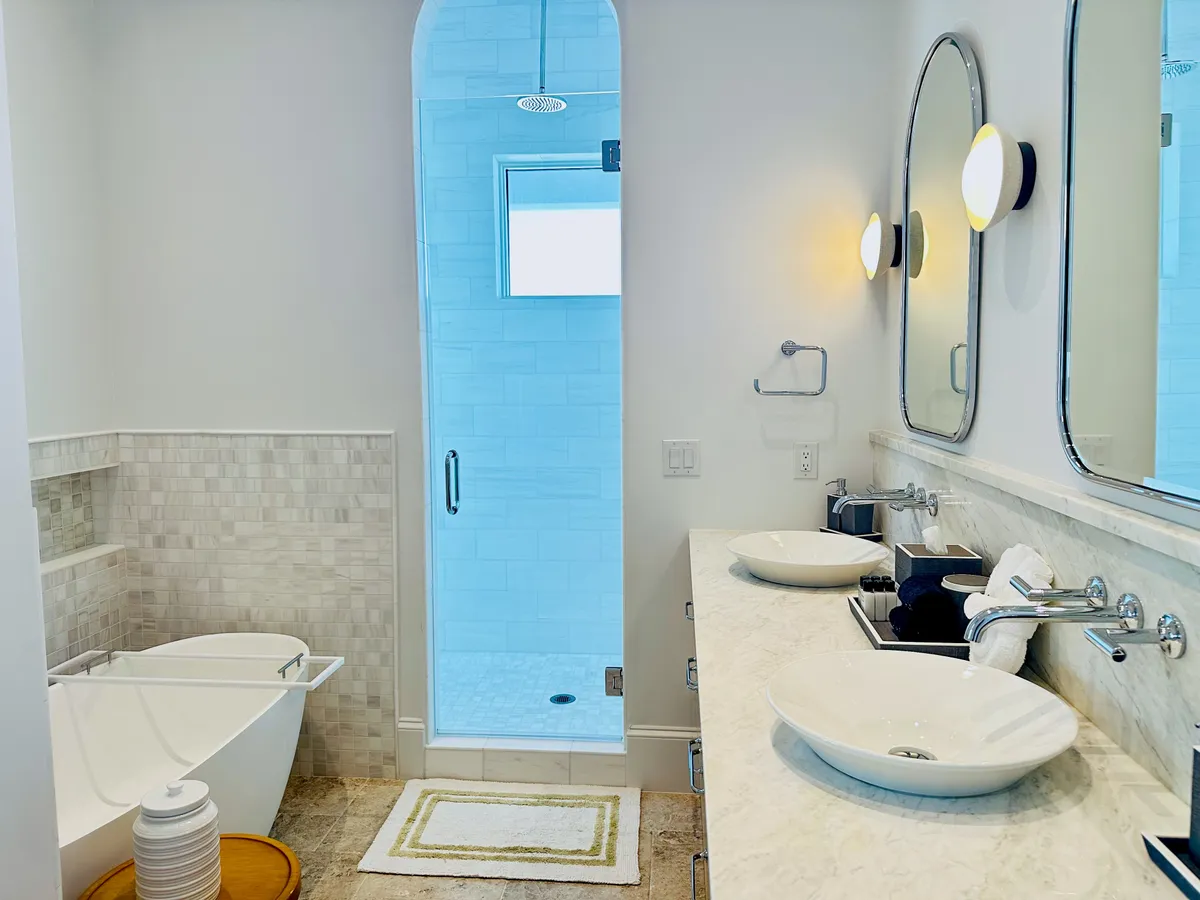 A bright, luxurious bathroom with a freestanding bathtub, glass-enclosed shower, double vessel sinks, and arched mirrors accented by wall sconces. The space exudes coastal elegance and offers a spa-like experience.