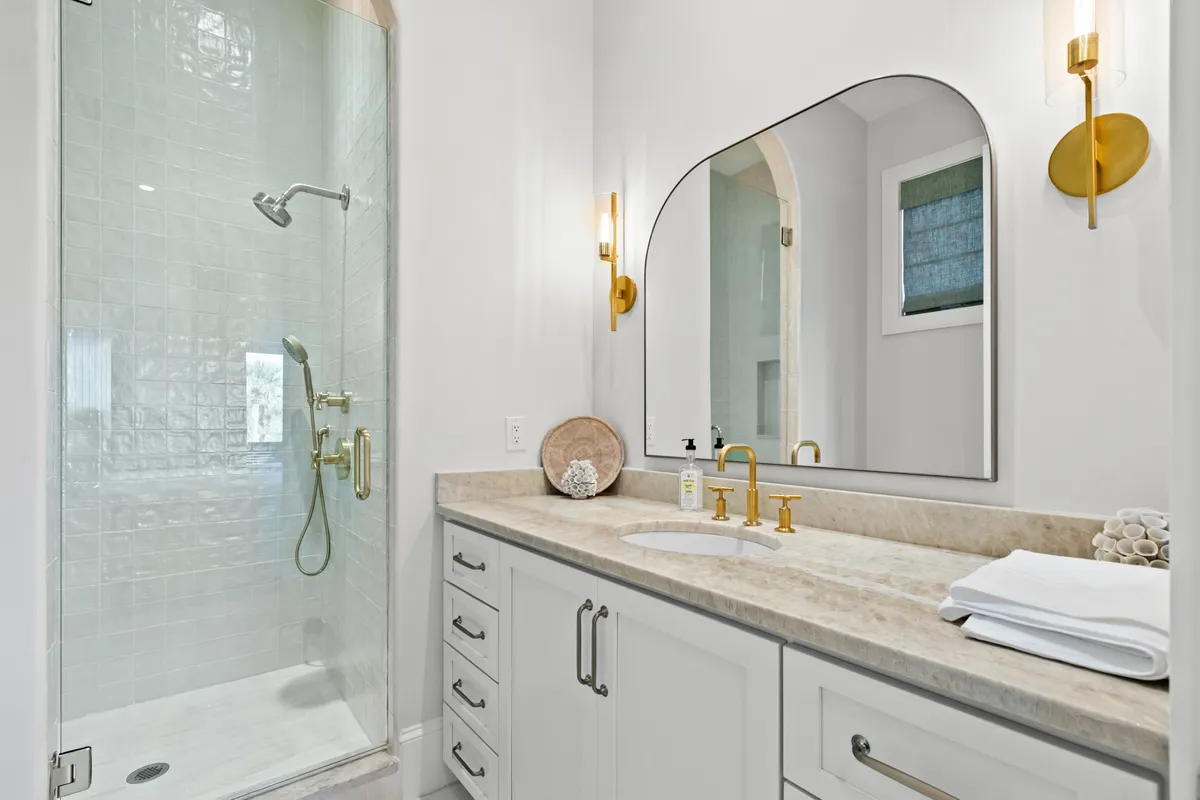 A stylish en-suite bathroom featuring a large arched mirror, glass-enclosed shower, gold fixtures, and a marble countertop. The space is designed for personal grooming and rejuvenation.