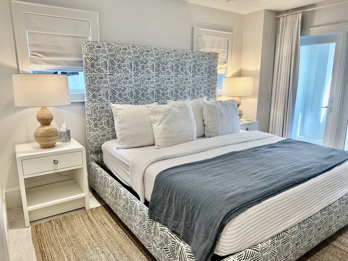 A cozy bedroom with a patterned queen bed frame, soft layered bedding and neutral pillows, matching bedside tables with lamps, and a glass door leading to a balcony or exterior. The overall feel is peaceful and inviting, perfect for relaxation.