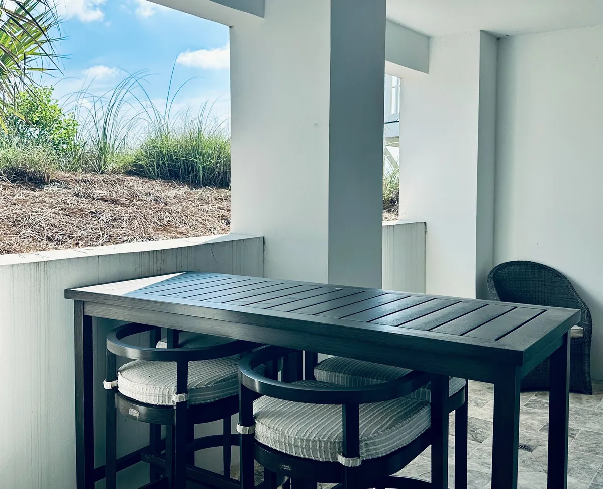 A stylish dining area featuring a dark wood table with cushioned stools, a large open window framing beach grass and blue sky, white walls and columns, and a cozy wicker chair in the corner. The space is filled with natural light, creating a relaxed yet sophisticated atmosphere for shared meals and conversation.