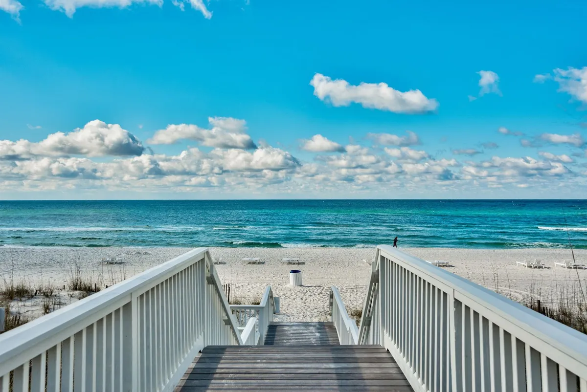 A white wooden boardwalk staircase descends onto a quiet, wide sandy beach with sparse vegetation. The vibrant blue ocean meets the horizon, and several lounge chairs are scattered along the shoreline, emphasizing tranquility and private enjoyment.