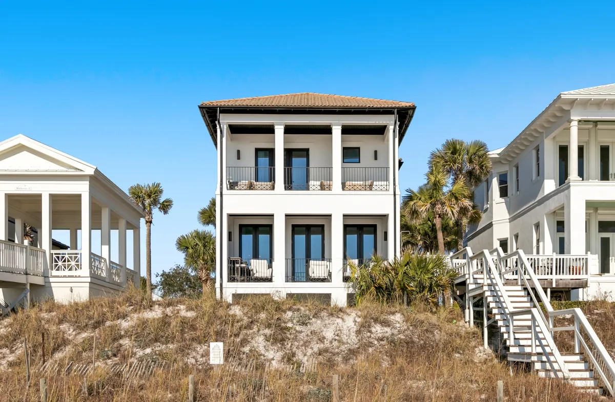 A three-story white coastal home with expansive balconies and large windows overlooks a natural dune landscape. A prominent staircase leads down to the sandy beach, with neighboring homes of similar coastal style nearby. The setting combines direct access to the beach with elevated outdoor living spaces.