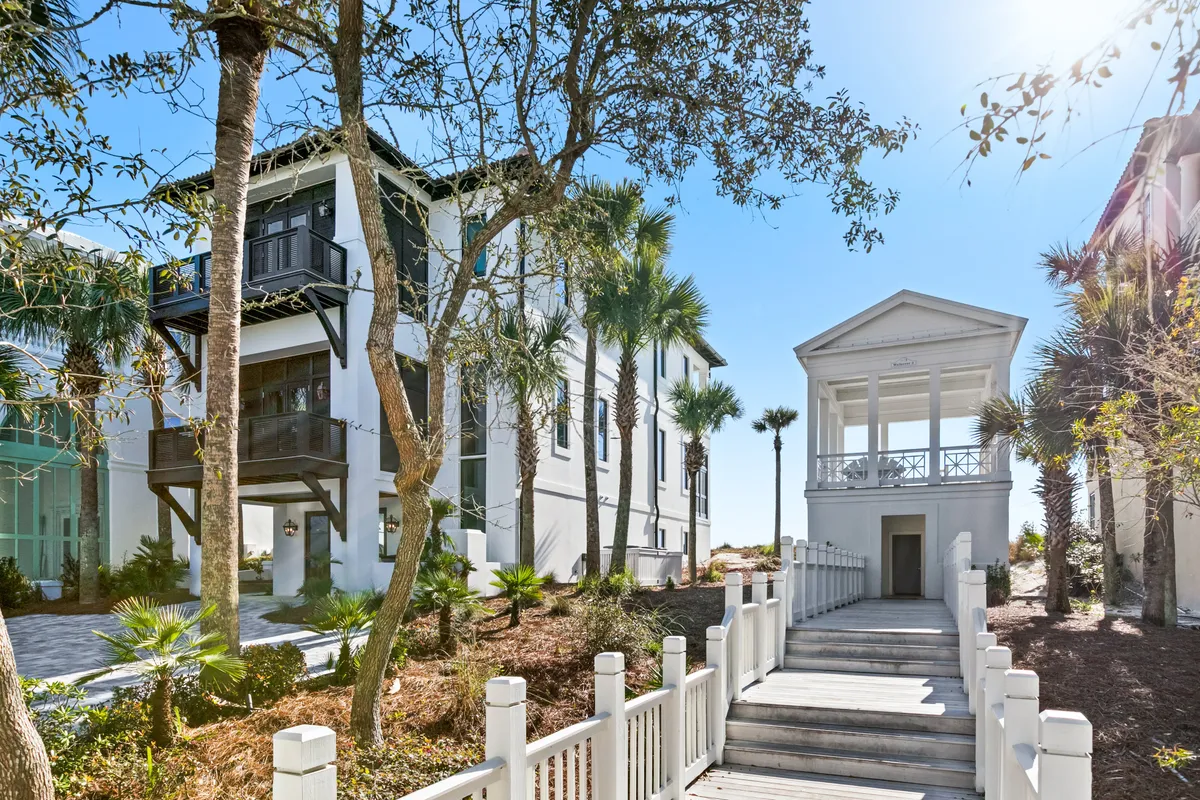 A picturesque white beach walkover with stately columns leads from a lushly landscaped home directly to the sandy beach. The wooden walkway is surrounded by palm trees and greenery, facilitating an easy, private transition from the residence to the shoreline.