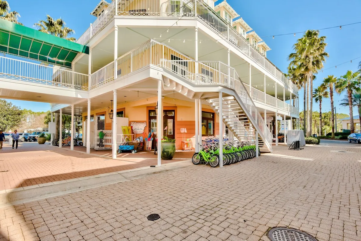 A sunlit courtyard with a row of green bicycles ready for guests, a multi-level peach and white building with balconies, palm trees, and string lights overhead. The paved courtyard feels lively and accessible, perfect for those who want to explore the neighborhood and nearby destinations by bike or on foot.
