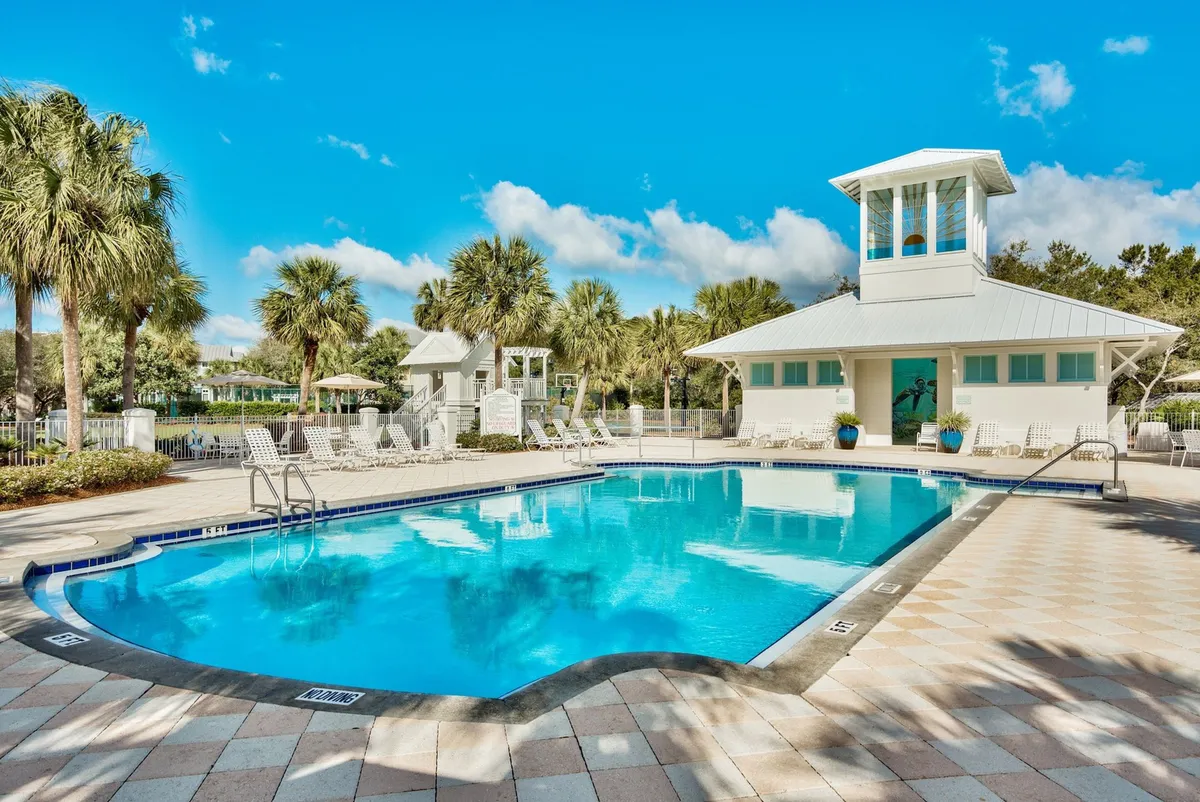 A sparkling blue community pool is surrounded by white lounge chairs and tall palm trees, with a distinctive white building featuring a belltower in the background. The area exudes a cheerful, welcoming atmosphere ideal for gatherings and leisure activities.