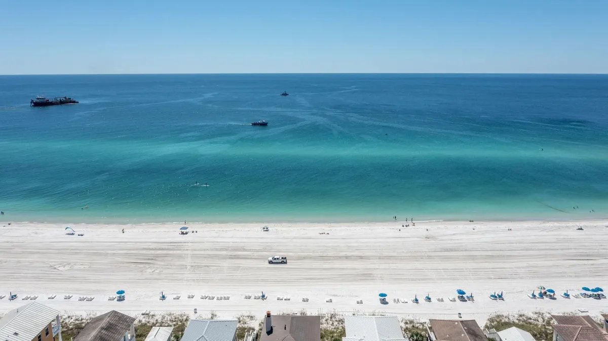 A pristine stretch of white sandy beach meets turquoise ocean waters under a clear sky. Rows of blue beach umbrellas and lounge chairs dot the shore, while boats float in the distance. The scene evokes peacefulness and exclusivity, offering a glimpse of serene evenings by the water and the allure of private, scenic beach access.