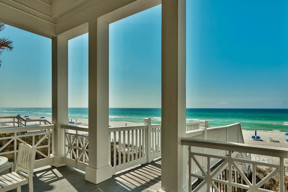 This image captures a white wooden boardwalk leading directly to a pristine, sandy beach with turquoise ocean waves. Several beach umbrellas are scattered across the sand, and a covered porch with white railing is visible in the foreground, offering a seamless transition from indoor comfort to the open beach.