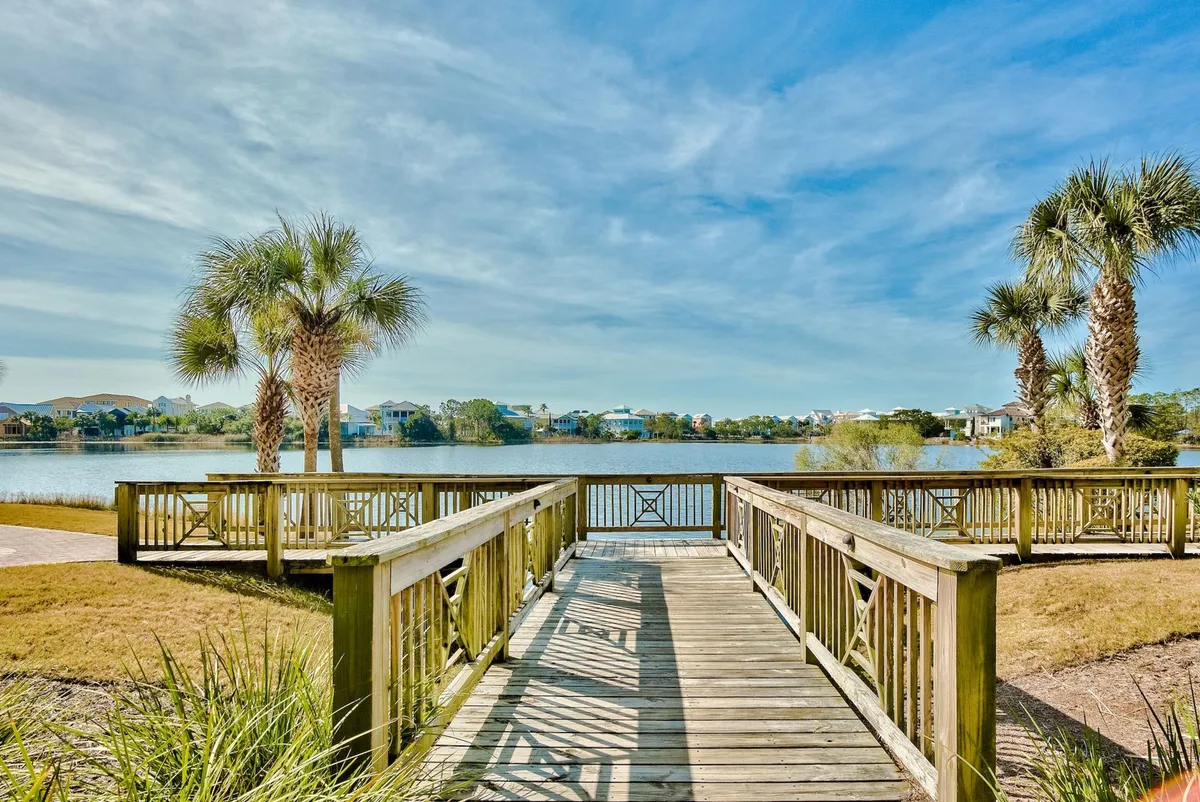 A wooden boardwalk with railings stretches over a calm water body, lined by palm trees and offering tranquil views of distant homes. The scene is serene and encourages family strolls, bird watching, and moments of relaxation in nature.