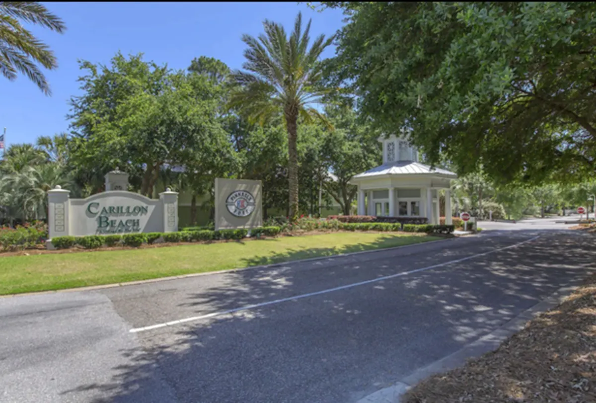 The welcoming Carillon Beach entrance sign and guardhouse set among palm trees, marking the official entry point into the neighborhood. The clean, palm-lined road evokes exclusivity and the anticipation of entering a beautiful, secure beach community.