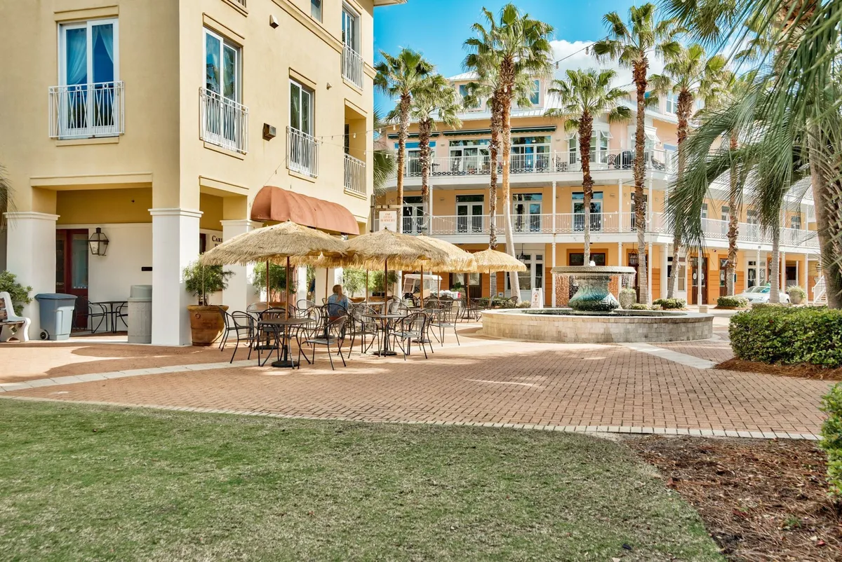 A central outdoor plaza featuring a charming fountain surrounded by lush palm trees, with inviting seating areas shaded by thatched umbrellas. The three-story building in the background, with balconies and large windows, hints at a vibrant, social neighborhood atmosphere. People can be imagined dining, socializing, and relaxing in this lively communal space.
